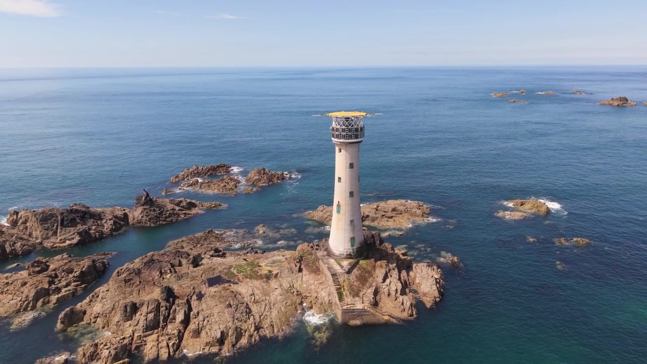 Faro di Hanois, Guernsey, Isole del Canale. Riprese ravvicinate e dettagliate con drone del faro su roccia di granito circondato da mare calmo e limpido e rocce sotto il sole del tardo pomeriggio con orizzonte nebbioso