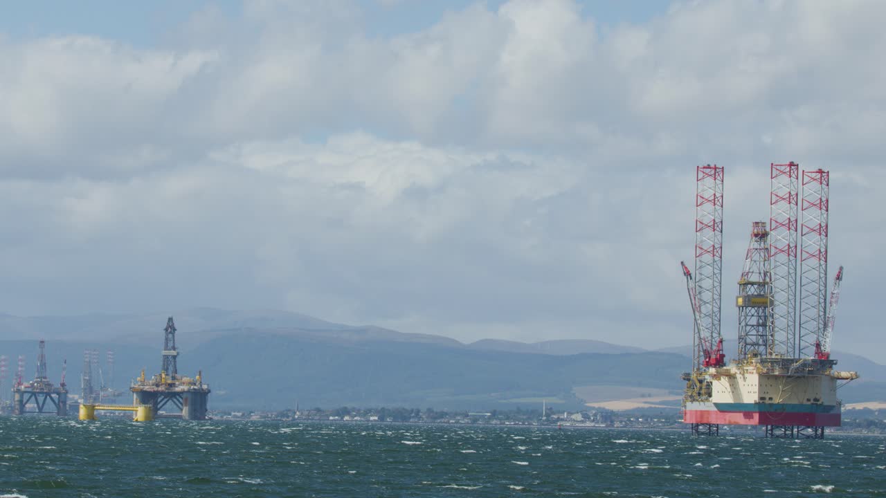 Two offshore oil rigs in rough sea under cloudy sky, distant hills and industry visible