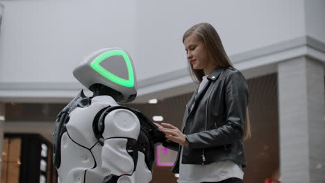 una chica sonriente con una camiseta blanca está en contacto con un humanoide. cyborg ayuda a la chica a responder preguntas. asistente robótico útil en la oficina y en el centro comercial