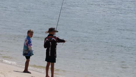 two people stand on a sandy beach, engaged in fishing by the water's edge.