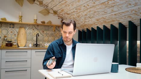 un joven ocupado con ropa casual de moda está hablando en el teléfono móvil y escribiendo en la computadora portátil en la cocina.