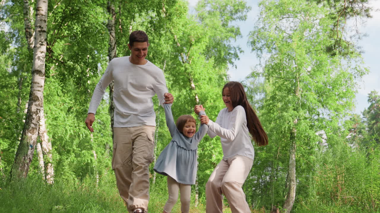 Happy children walking together on forest path holding hands, gently lifting youngest sibling between them, surrounded by green trees and sunlight, creating warm joyful outdoor family moment