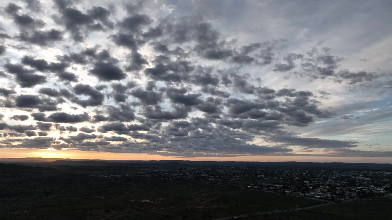 Elevated aerial establishing shot of Broken Hill beneath a dense textured cloudscape, with a narrow band of warm sunrise colour separating the dark landscape from the morning sky.