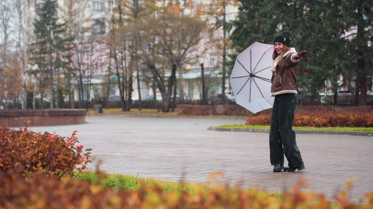 Lady holding umbrella, wearing knit cap, brown shearling jacket, black trousers, happily turning around in light snowfall, enjoying winter day, spreading hands, embracing cold weather joyfully