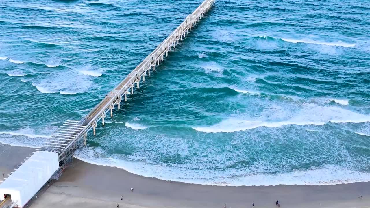 Drone footage showcasing a long pier extending into the ocean with waves crashing below.