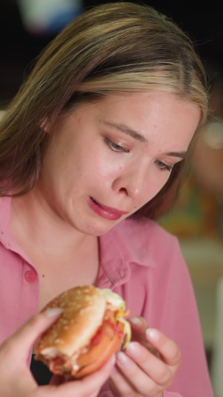dama de vestido rosa examina la hamburguesa con una expresión de descontento, mostrando signos de disgusto antes de dejarla caer, la gente en el fondo está comiendo, con la vista borrosa y las luces iluminando suavemente