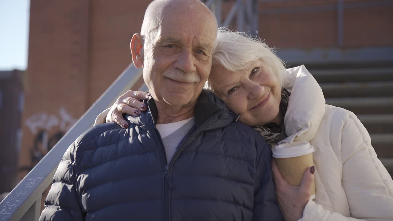vista cercana de una pareja mayor abrazándose y mirando la cámara en la calle en un día de invierno. la mujer sostiene un café para llevar