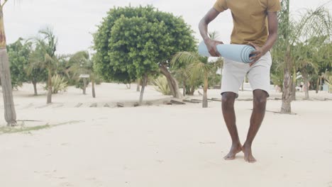 happy african american man rolling up mat after doing yoga at the beach, slow motion