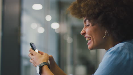 Young-Businesswoman-Sitting-On-Floor-In-Corridor-Of-Modern-Office-With-Phone-Celebrating-Good-News