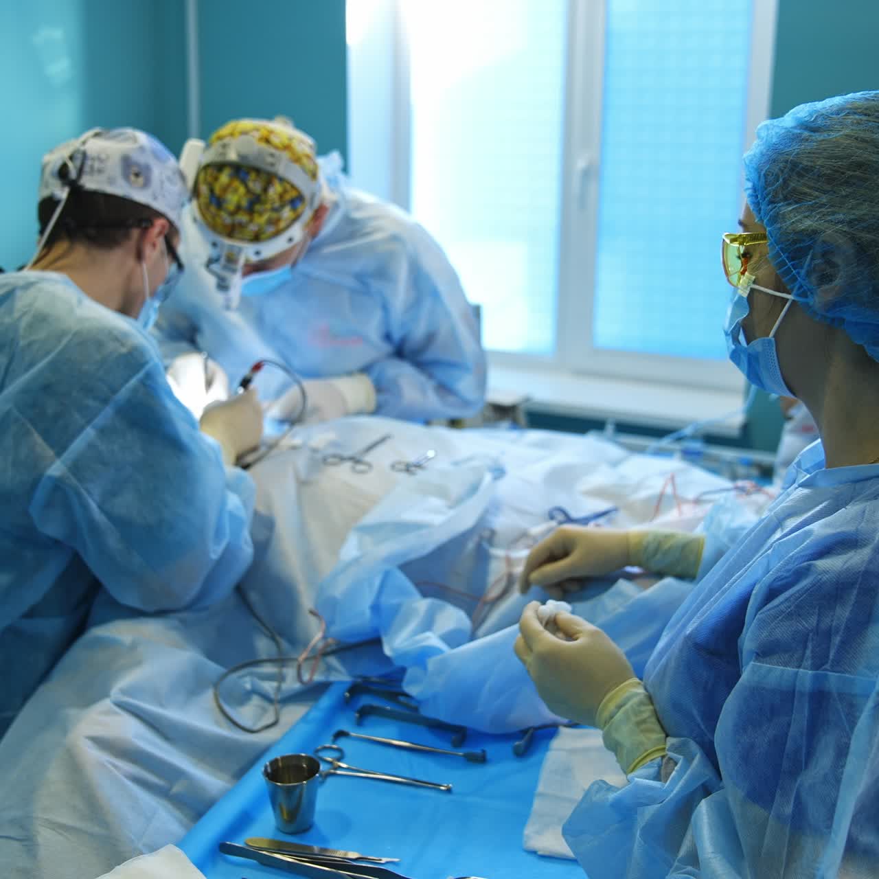 Surgical nurse standing near the table with tools and instruments looking at surgery performed by two doctors. Side view