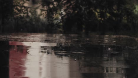 Rainfall-with-puddles-on-a-footpath-in-the-background-the-road-and-a-passing-car-behind-the-bushes---close-up-shot