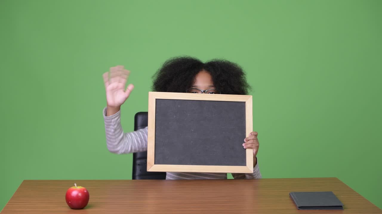 Giovane ragazza africana carina con capelli afro che mostra una lavagna mentre è seduta