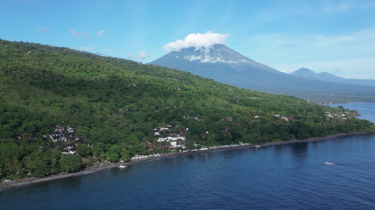 Sunny blue skies in Amed, Bali with views of Mount Agung covered in clouds and sea in the foreground, aerial
