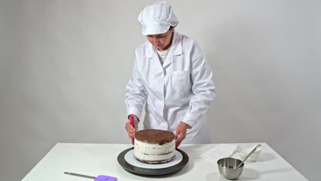 baker removes the excess cream from the sides of a chocolate cake by rotating the stand and using a spatula.