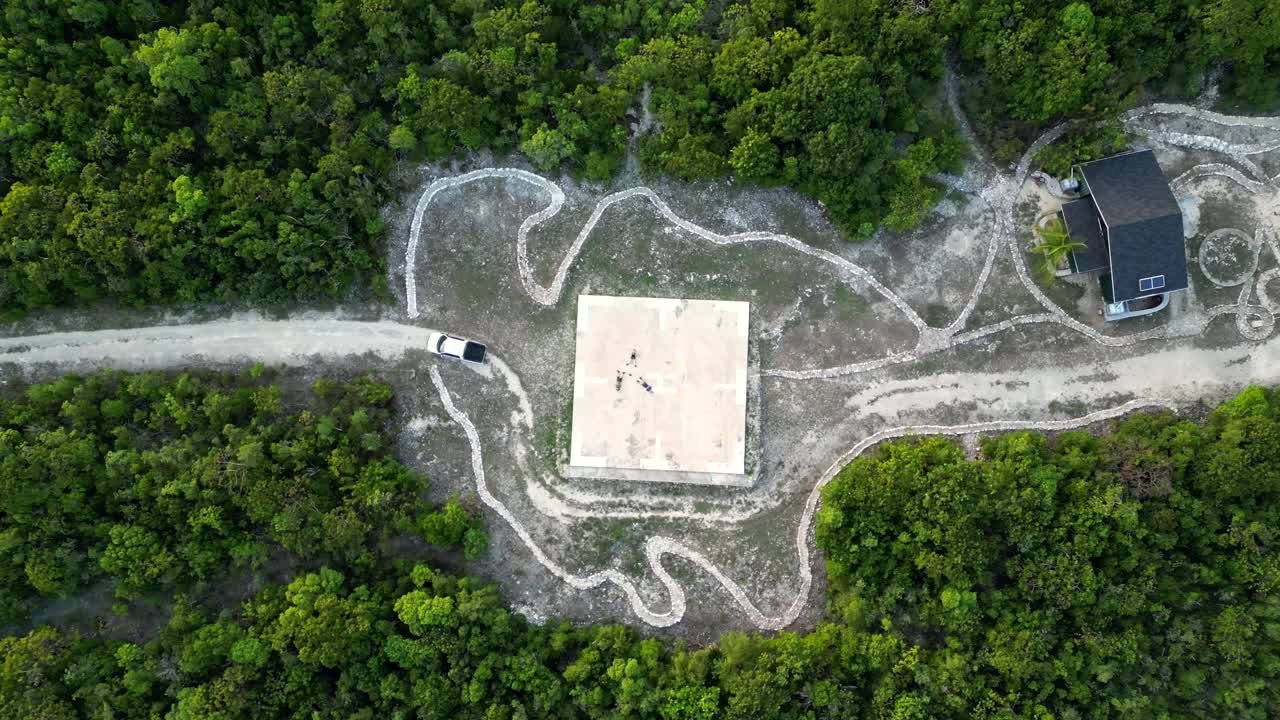 드론이 천천히 트럭과 카리브해의 케이먼 섬의 열대 우림 한가운데에 있는 헬리 ⁇ 터 기지 위에 누워 있는 세 명을 향해 내려갑니다.