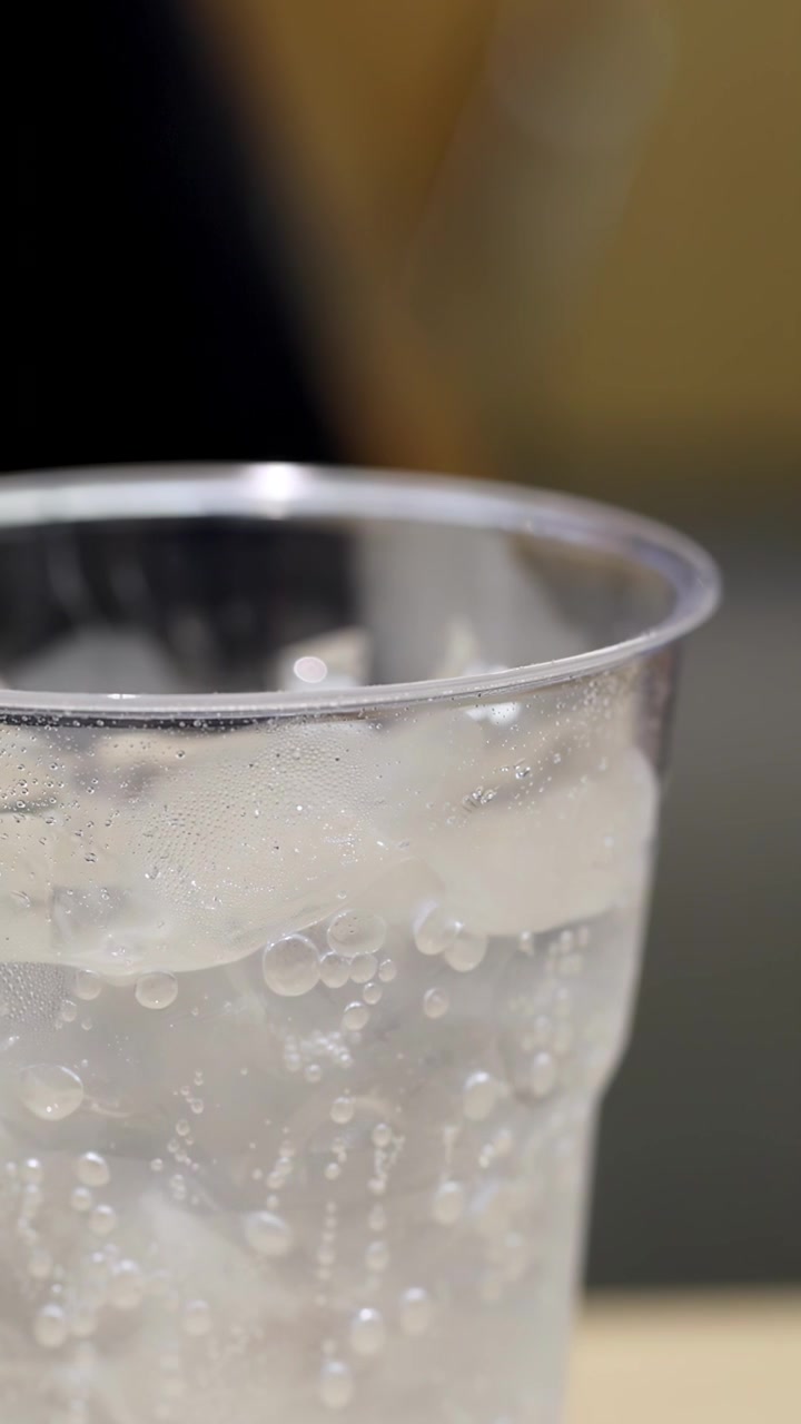 柔らかな照明の下、プラスチックカップに入った氷入り炭酸飲料のマクロビュー。泡が立ち上る。