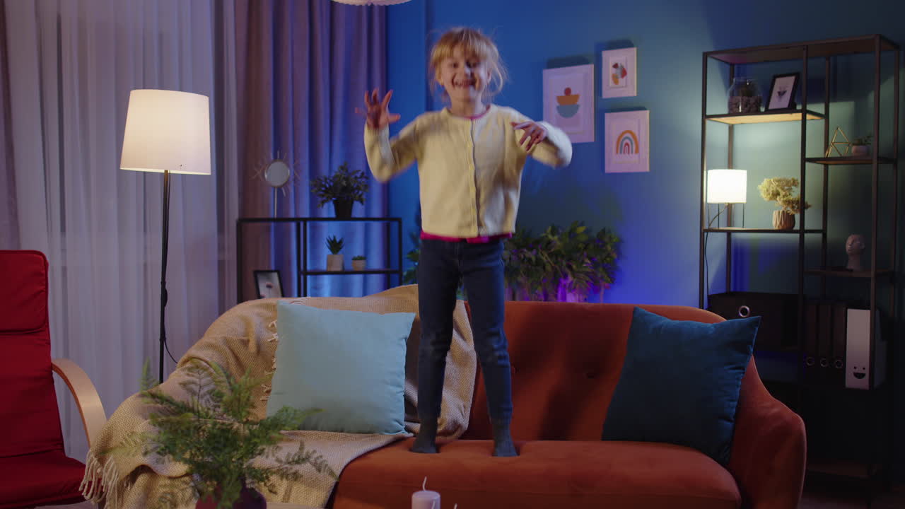 Happy child girl kid dancing trendy dance while listening to music at home alone standing on sofa