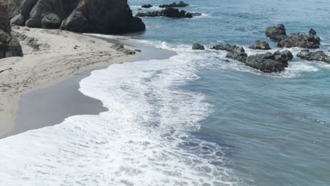 Low-altitude-aerial-up-tilt,-over-waves-crashing-on-empty-beach-by-Little-Sur-River-Beach-near-highway-1-in-California,USA,-revealing-huge-rocks-and-cliffs-on-the-coast-and-in-the-water