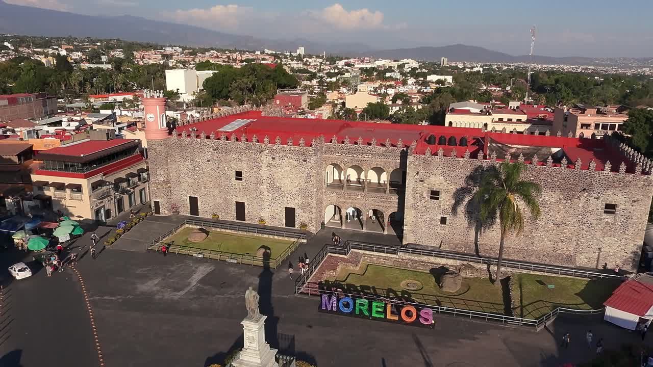 Overhead drone view of Cuernavaca’s city center showing Palacio de Cortés, Carlos Pacheco statue, palm tree, and vibrant Morelos letters in bright daylight.