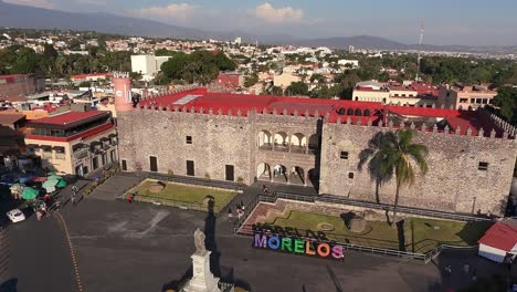 overhead drone view of cuernavaca’s city center showing palacio de cortés, carlos pacheco statue, palm tree, and vibrant morelos letters in bright daylight.