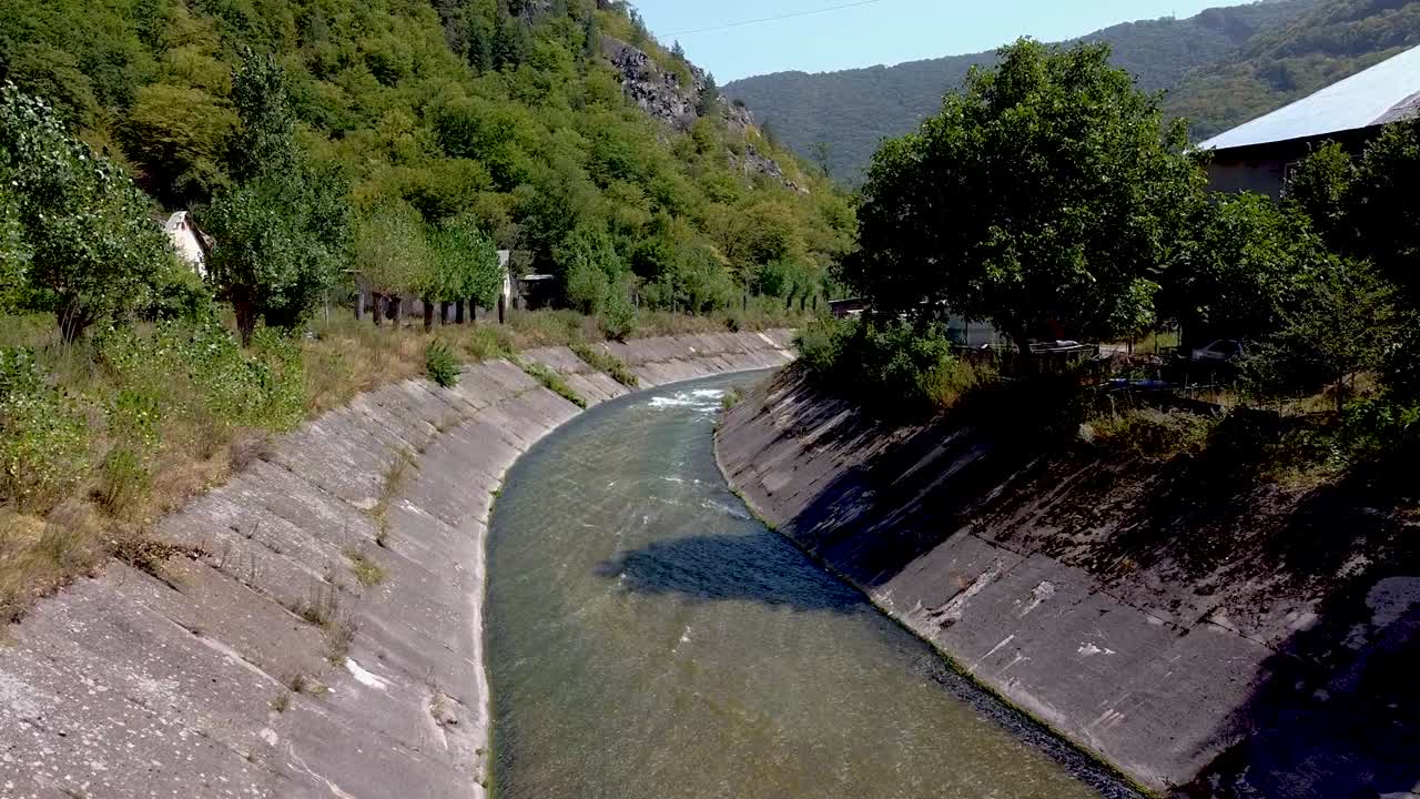 A clear river gently runs through a valley surrounded by hills and trees. The calm water reflects the lush greenery, creating a peaceful natural setting