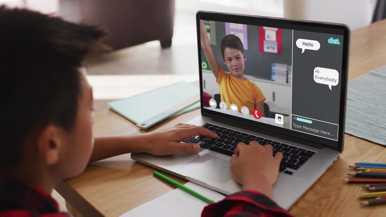 Schoolboy using laptop for online lesson at home, with boy raising hand and webchat on screen