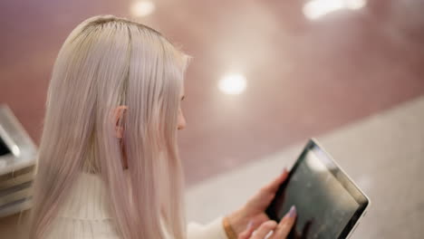 high angle view of internet user seated on bench using manicured nails to tap glossy tablet screen, blurred mall floor background and subtle elegant lighting highlighting modern browsing gesture