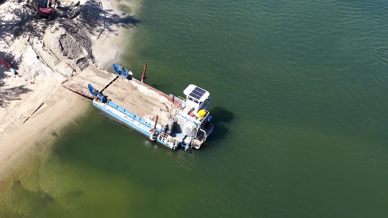 Filmato drone dall'alto mostra macchinari industriali che effettuano il ripascimento della spiaggia lungo un litorale soleggiato.