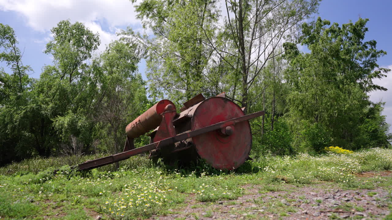 Rusty agricultural equipment lies abandoned in a grassy field surrounded by trees and flowers on a sunny day