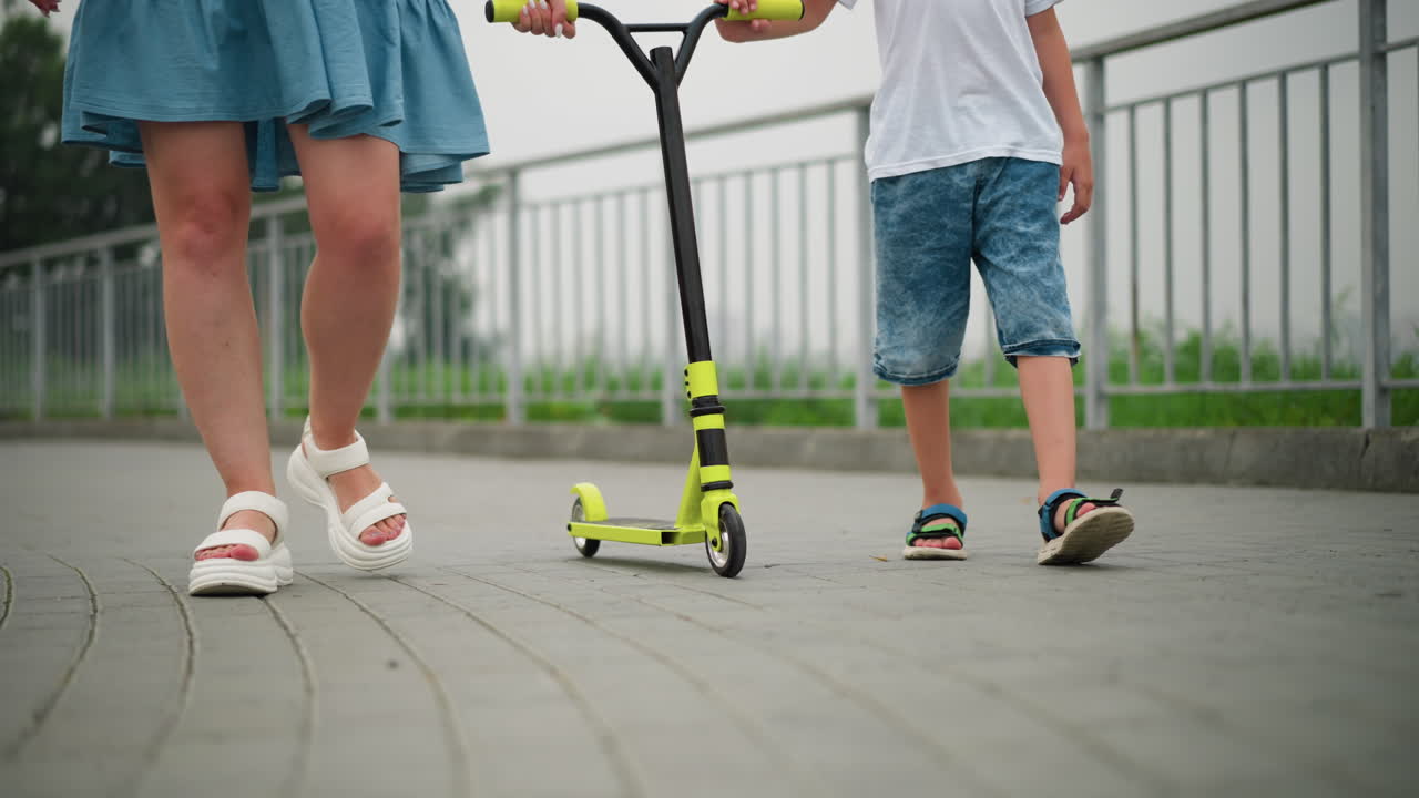 A lower view of a woman and a child walking on a paved walkway, pushing a yellow scooter, both are dressed casually, with the focus on their legs and the scooter