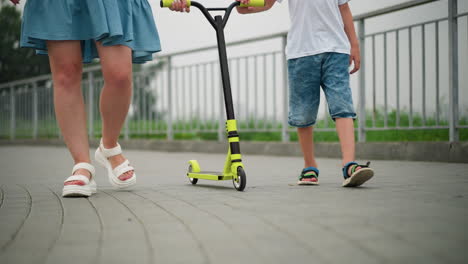 a lower view of a woman and a child walking on a paved walkway, pushing a yellow scooter, both are dressed casually, with the focus on their legs and the scooter