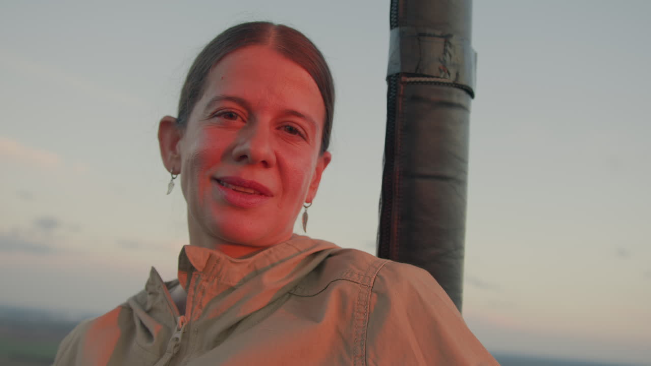 Confident woman with subtle smile looks ahead while resting beside vertical structure in open-air setting, warm sunset light gently highlighting face