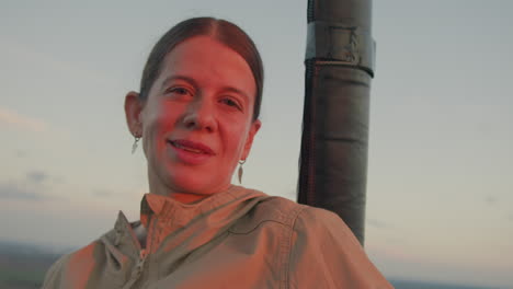 confident woman with subtle smile looks ahead while resting beside vertical structure in open-air setting, warm sunset light gently highlighting face