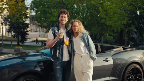 a guy and a girl are standing near a gray cabriolet smiling and looking at the camera and drinking yellow cocktails