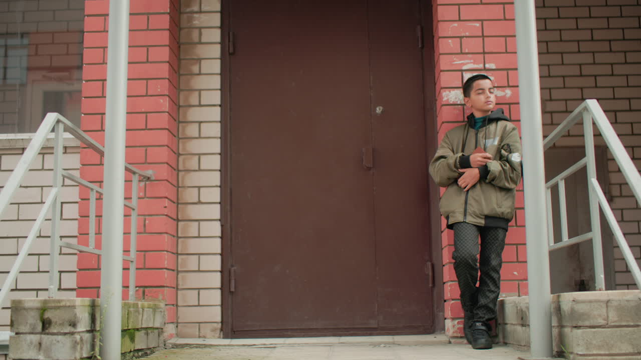 Boy in hooded jacket leans against red brick wall near building entrance holding brown book, gazing thoughtfully around with calm expression in quiet urban outdoor environment