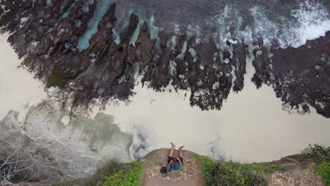 Tourist-person-sitting-on-a-steep-cliff-edge-of-Broken-Beach-on-Nusa-Penida-Island,-looking-at-waves-breaking-through-a-natural-arch-bridge-in-the-horizon