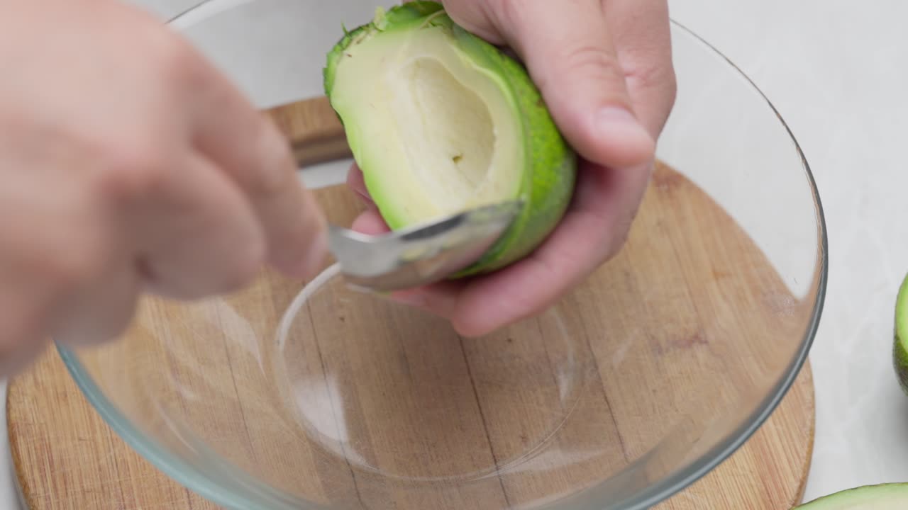 Primo piano della polpa di avocado che viene prelevata con un cucchiaio e messa in una ciotola trasparente su una tavola di legno
