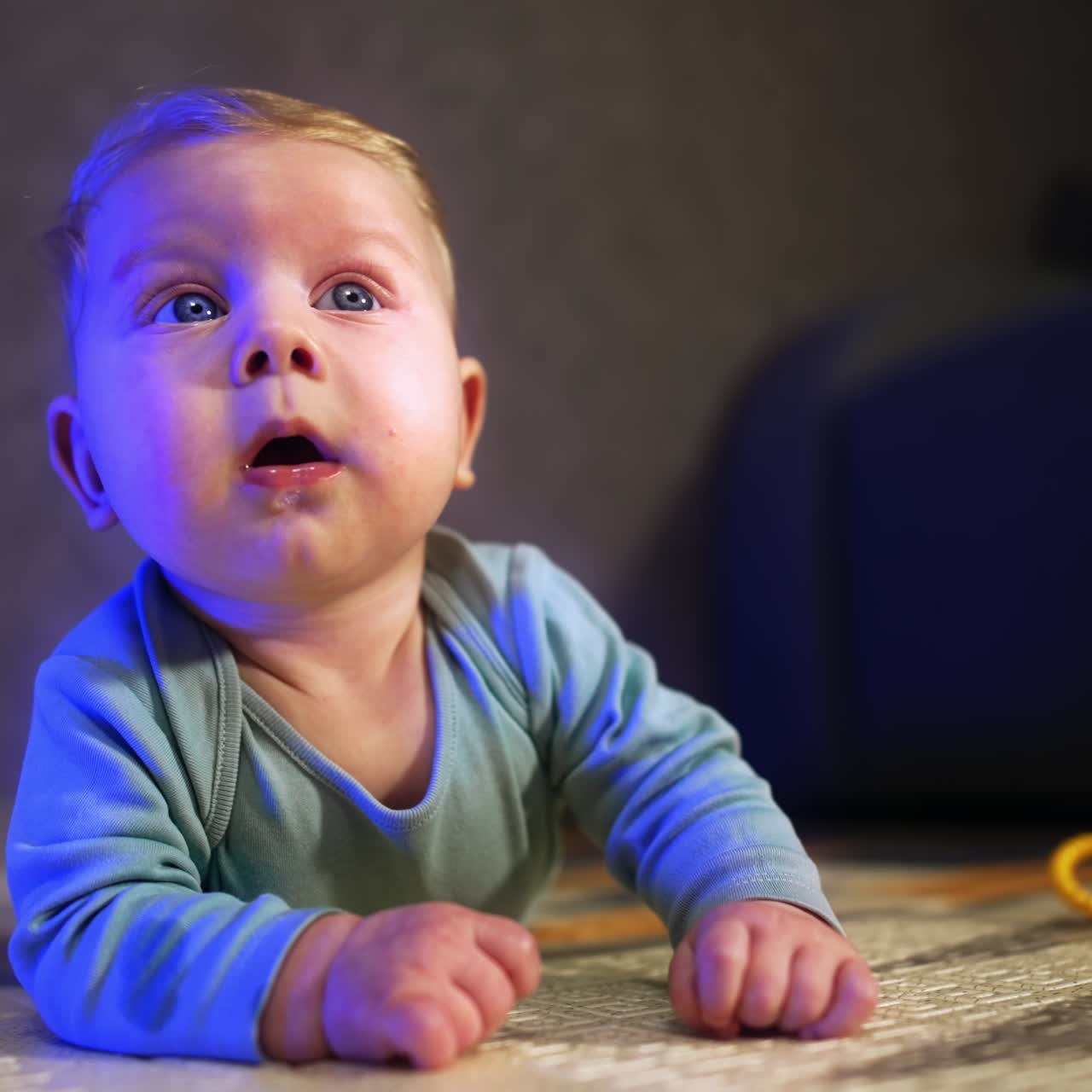 Søt, kaukasisk baby med blå øyne ligger på magen. Nysgjerrig liten gutt som ser seg rundt. Uskarp bakgrunn