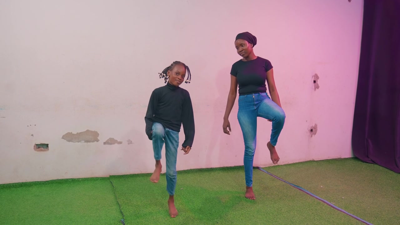 Madre e figlio neri imparano passi di danza su erba sintetica interna con parete rosa, madre che funge da mentore e allenatrice, postura concentrata e sollevamenti coordinati delle gambe, prova giocosa che costruisce fiducia e legame
