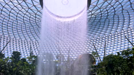 a large indoor waterfall pours from a circular skylight into a lush garden atrium, surrounded by greenery and visitors, under bright natural daylight
