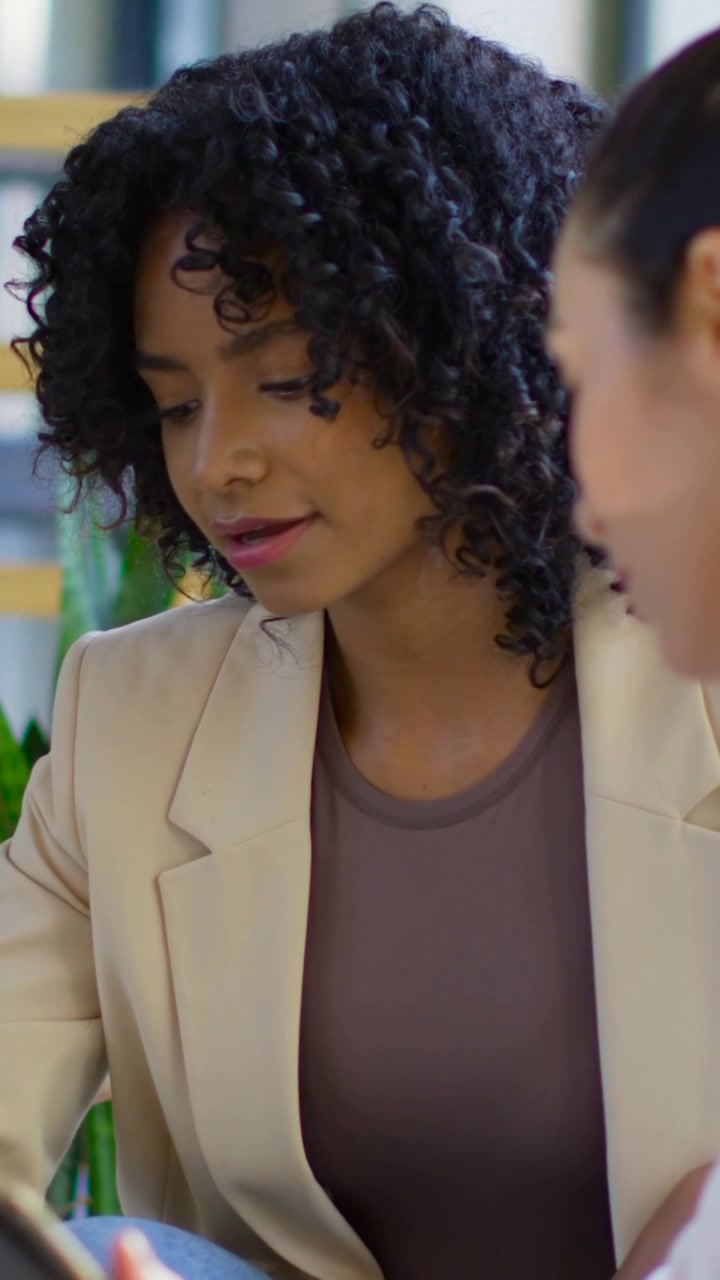 Vídeo vertical: Mulher de top branco trazendo o telefone, mostrando o projeto, enquanto uma pessoa de blazer bege revisa o escritório.