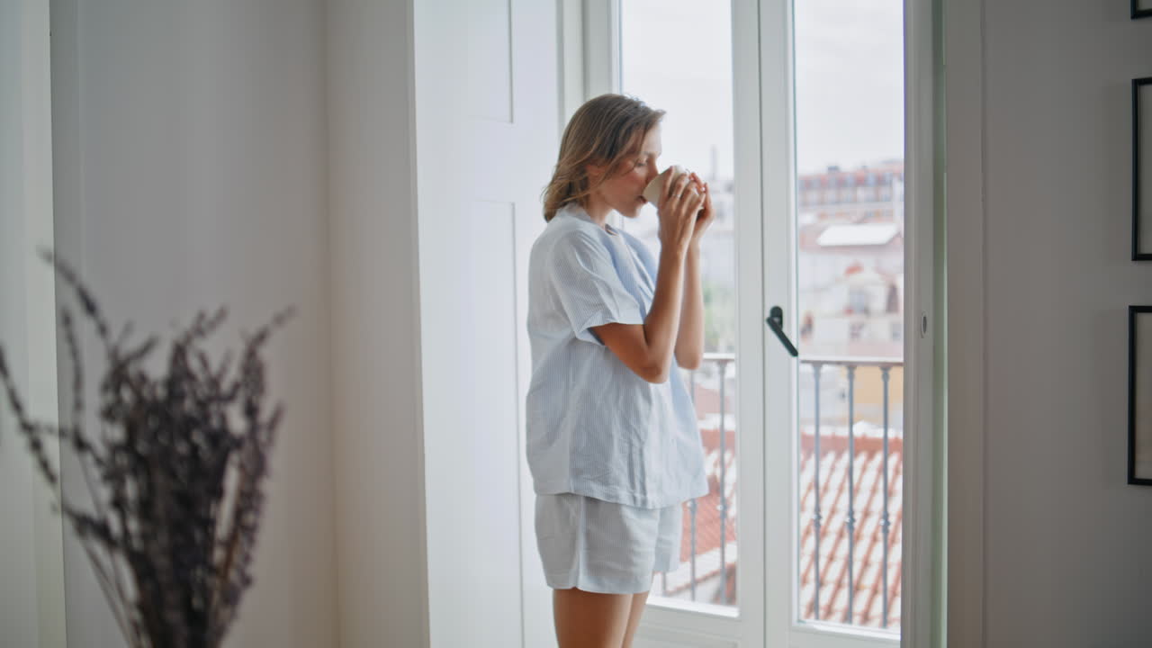 Ragazza si gode il caffè del mattino in un appartamento accogliente. Donna rilassata beve qualcosa indossando abiti comodi