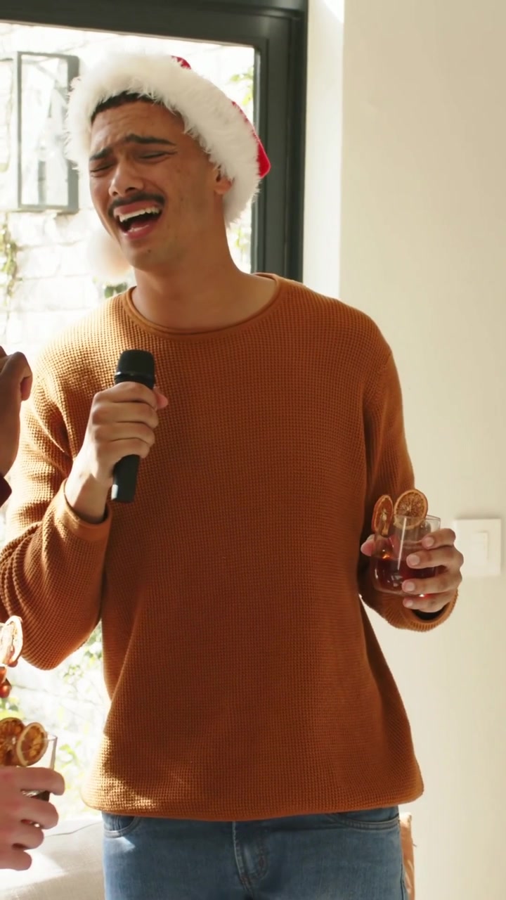Vertical video: Cue Santa-hat man in sweater singing holding mic and cocktail celebrating at home
