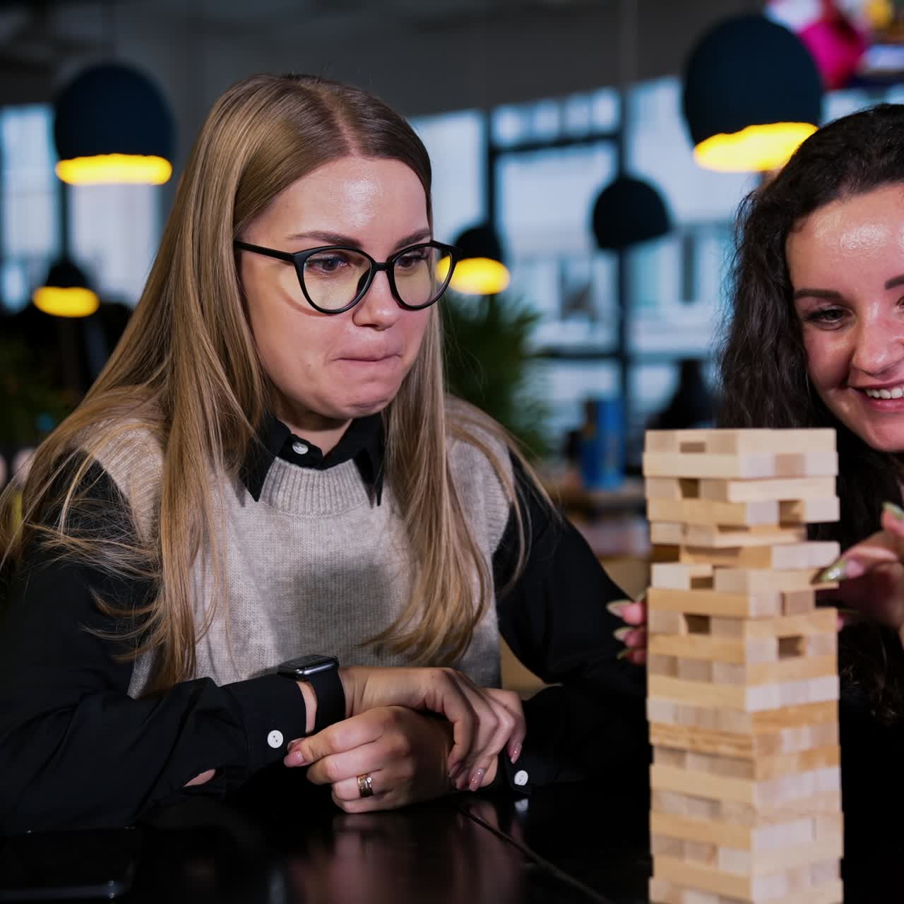 Colleagues having a break from work playing board games. Two ladies pull the wooden brick from a pile in jenga game