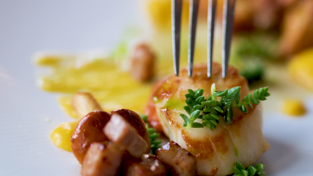A fork lifts a seared scallop garnished with herbs from an artfully plated seafood dish in a fine dining setting, under soft natural lighting