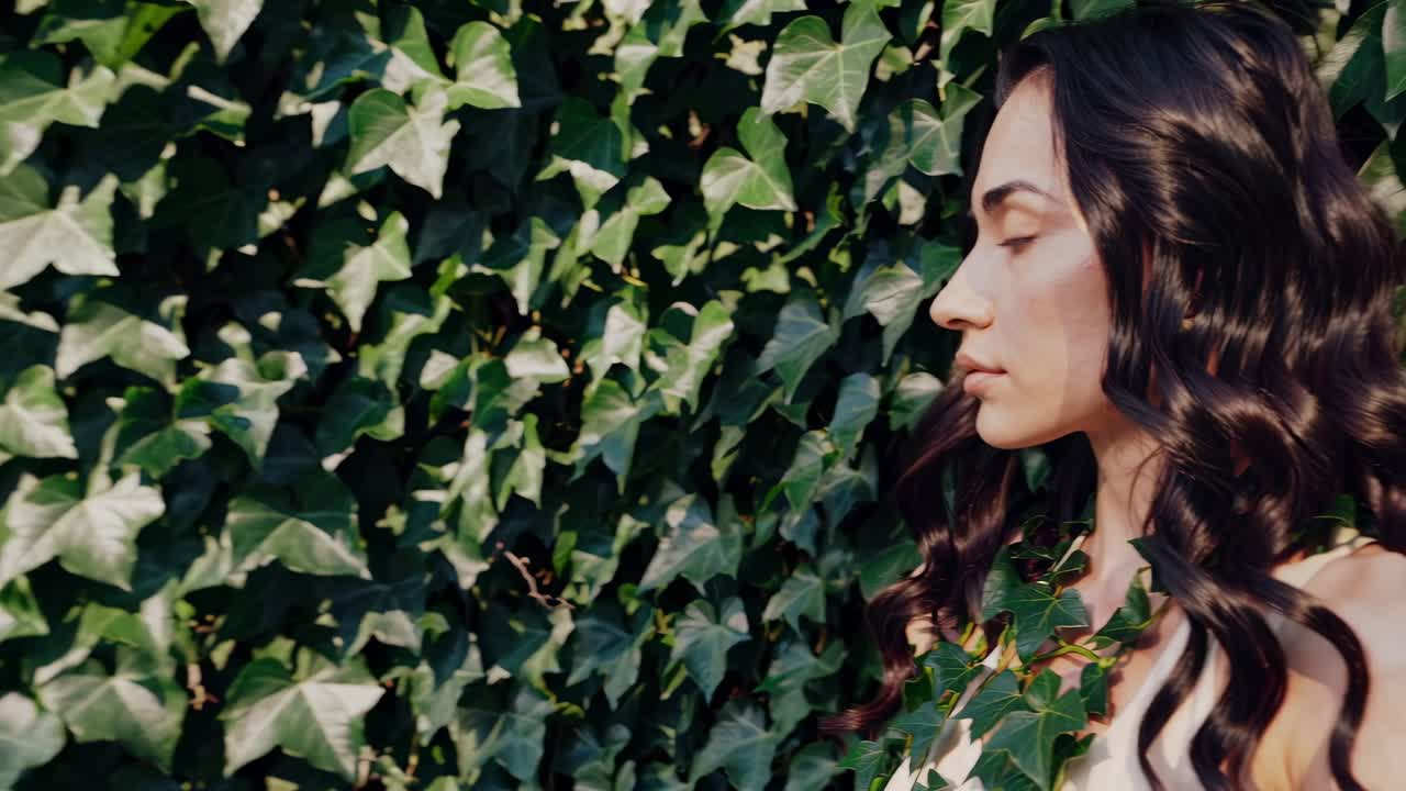 Woman with long, flowing hair stands peacefully against a vibrant wall of ivy. Her eyes are closed, suggesting a moment of tranquility and connection with nature