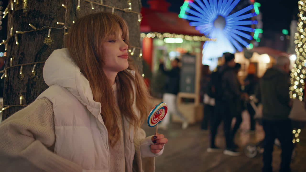 Pretty lady licking lollipop looking smartphone at night luna park closeup.