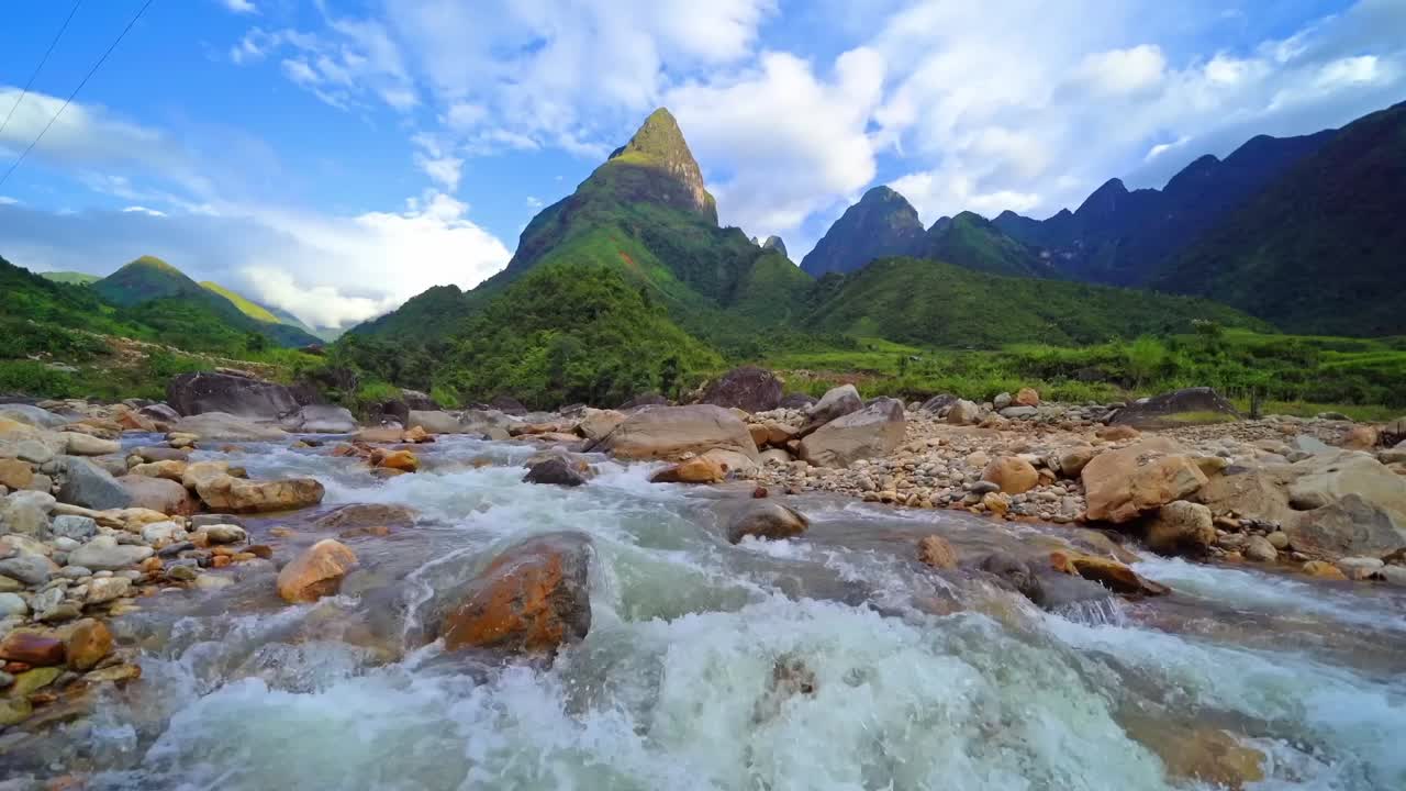 A fast-flowing mountain river with clear whitewater rushes over rocks beneath a towering green peak under a blue sky with scattered clouds in a remote valley.