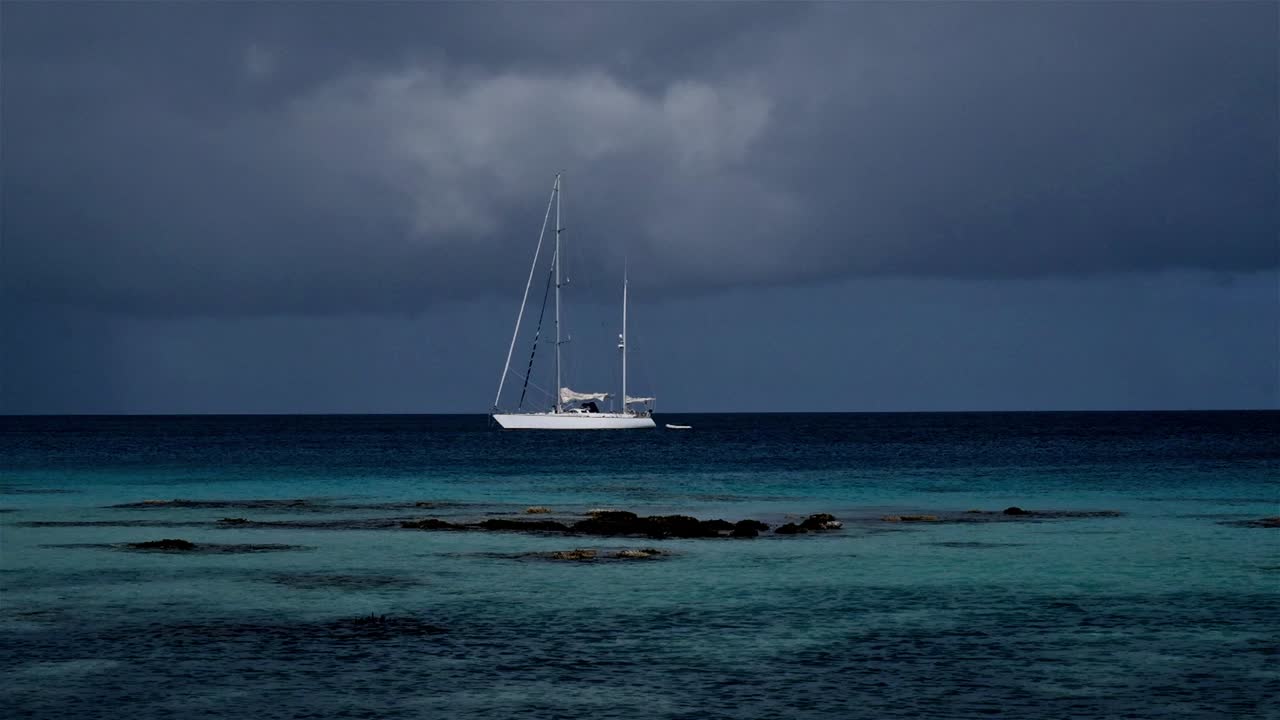 un yate de vela fondeado en un paraíso tropical con nubes de tormenta en la distancia y un arrecife poco profundo delante