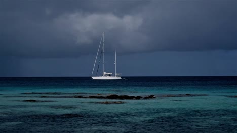Sailing-yacht-at-anchor-in-a-tropical-paradise-with-storm-clouds-brewing-in-the-distance-and-a-shallow-reef-in-front